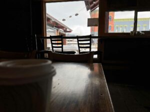 Interior view of Buchi Cafe Cubano in Leadville, Colorado with a to-go coffee cup on the table, two silhouettes of chairs, and colorful historical buildings across the street.