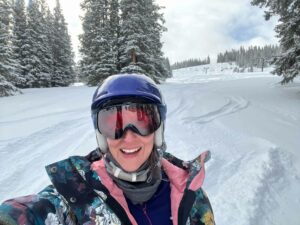 White woman snowboarder in her 40s at Ski Cooper wearing a purple ski helmet, goggles, and a floral coat with a background of trees and powder