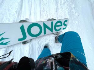 A Jones snowboard turned upside down and the legs of a white woman snowboarder in her 40s wearing electric blue snow pants buried in the snow when she got stuck.