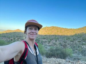 white woman in her 40s wearing a baseball cap taking a selfie at Saguaro National Park with the desert landscape in the background