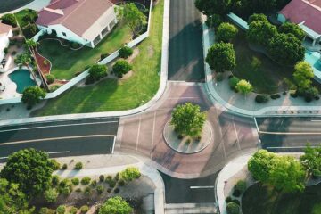 Aerial view of a quiet suburban neighborhood with curving streets, landscaped yards, and houses featuring red tile roofs. The image shows a small roundabout at the intersection of three roads, surrounded by green lawns, trees, and fenced backyards—some with swimming pools. The setting appears to be in a warm, sunny climate.